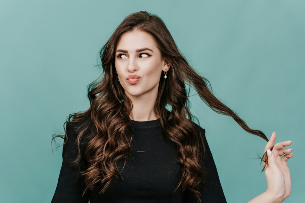 Playful woman in sweater looking aside touches hair looking aside over studio turquoise background